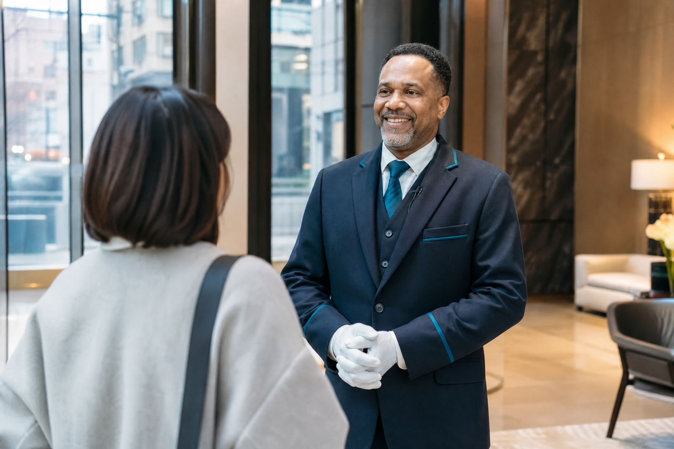 A professional doorman in a navy uniform with teal accents greets a resident in a modern NYC high-rise lobby.