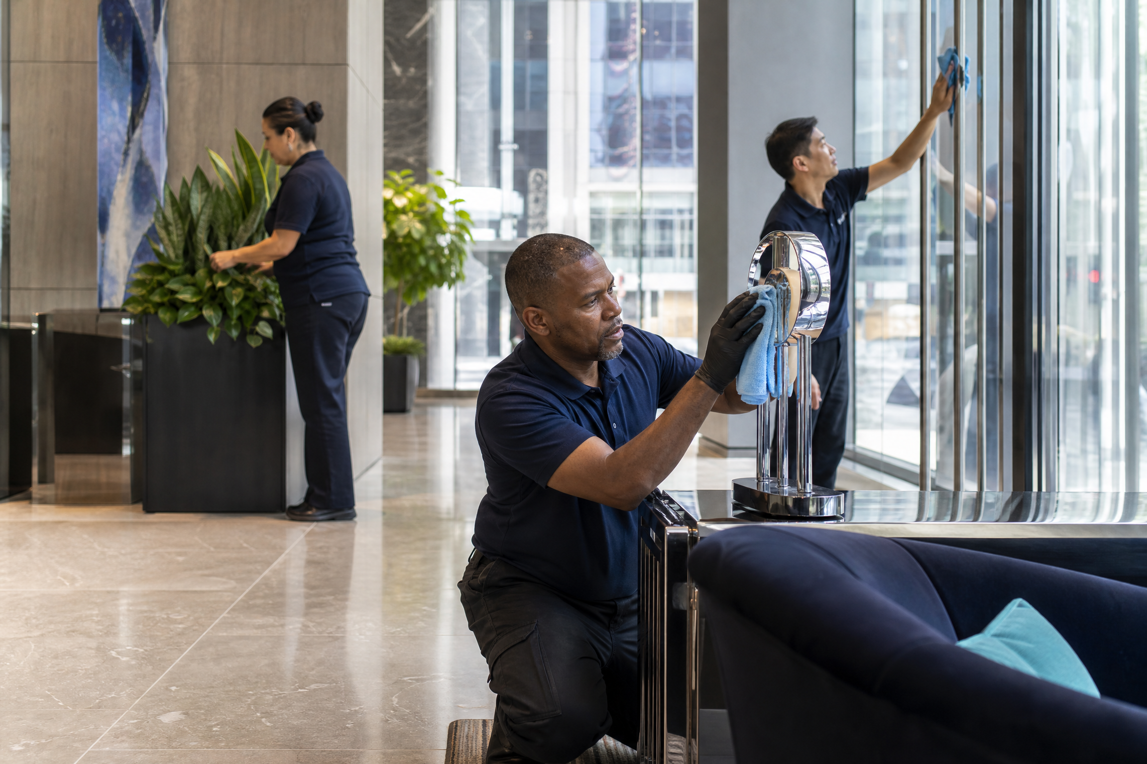 A diverse team of porters in a modern New York City high-rise lobby, polishing fixtures, arranging plants, and cleaning glass, with natural light and navy/teal accents.